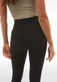 Leggings neri a vita alta realizzati in tessuto elastico, con cuciture curve, due tasche posteriori e una texture liscia.