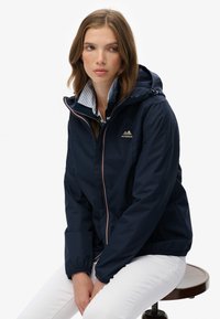 Jeune femme aux cheveux châtain clair portant une veste à capuche bleu marine et un pantalon blanc, assise sur un tabouret en bois, regardant sur le côté.