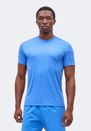 Homme debout, face à l’objectif, portant un t-shirt de sport bleu vif à manches courtes et un short bleu assorti sur un fond uni.