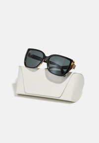 Ochelari de soare mari, negri și cu model țestoasă, cu lentile întunecate, expuși pe o carcasă albă cu textură, gravată cu "MICHAEL KORS."