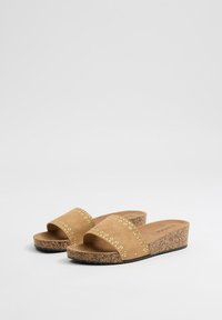 Sandalias deslizantes de ante marrón con acentos de ojales dorados, suela de corcho y diseño de punta abierta. Base plana para comodidad y estabilidad.