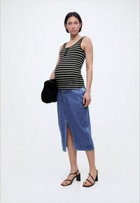 Rase naine, kes kannab musta-valget triibulist t-särki, midi pikkust teksaseelikuga, millel on esikäik, musti sandaale ning hoiab valgel taustal musta jopet.