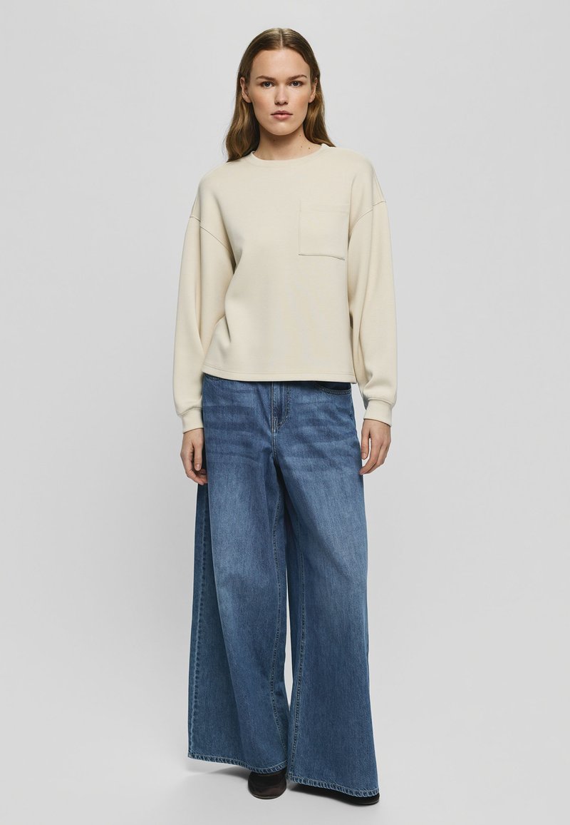 Femme aux cheveux lisses portant un haut crème à manches longues et un jean bleu à jambes larges, debout devant un fond gris clair uni.
