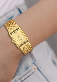 Kuldne ristkülikukujuline käekell, millel on tekstuuriga kett ja rooma numbrite märgid. Omab punutud metallist rihma ja stiilset, minimalistlikku disaini.