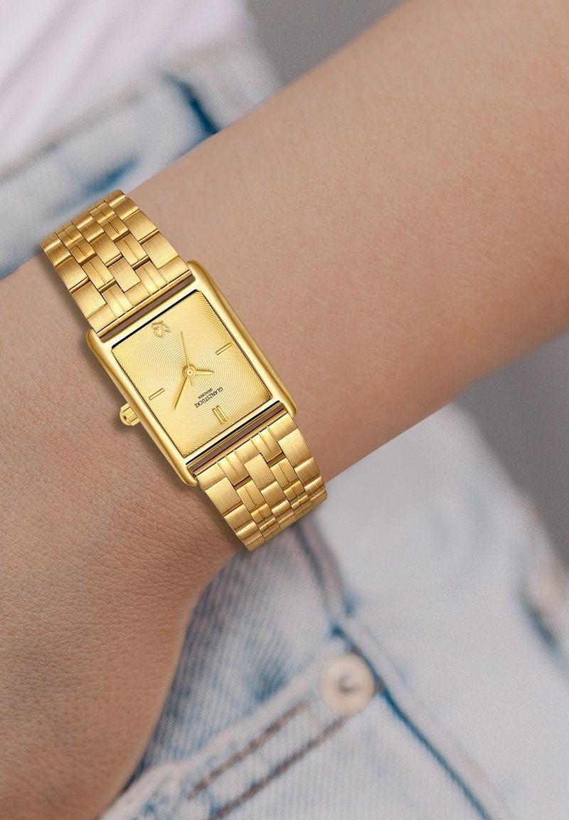 Kuldne ristkülikukujuline käekell, millel on tekstuuriga kett ja rooma numbrite märgid. Omab punutud metallist rihma ja stiilset, minimalistlikku disaini.