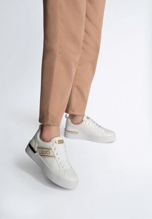 Person trägt beigefarbene Hose und weiße Plateausneaker mit goldenen Akzenten und Markenlogo vor weißem Hintergrund.