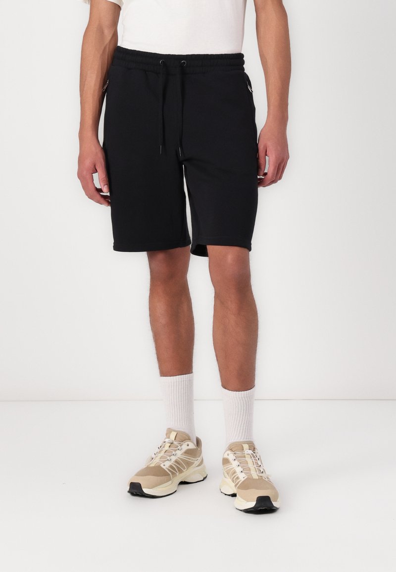 Shorts de sport noirs avec une ceinture à cordon de serrage ajustable, des poches latérales et une texture douce, associés à des baskets beiges et des chaussettes blanches.