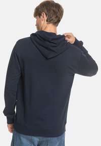 Tmavě modrá mikina s kapucí na šňůrku a žebrovanými manžetami. Vyrobena z měkké tkaniny, s uvolněným střihem a jednoduchým designem.