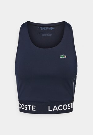 Granatowy, bezrękawnikowy crop top Lacoste Sport z białym elastycznym obszyciem z marką "Lacoste" i zielonym logo krokodyla na piersi.