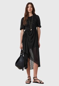Vestido negro transparente con lunares texturizados, escote fruncido, dobladillo asimétrico, combinado con un bolso negro y sandalias de tiras. Incluye un collar largo.