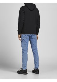 Persona che indossa una felpa con cappuccio nera e jeans azzurri chiari con molte tasche, in piedi contro uno sfondo neutro, di spalle.