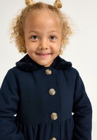 Bambino con capelli biondi e ricci in uno chignon spaziale, indossa un cappotto blu navy con grandi bottoni e un colletto di pelliccia, sorridente.