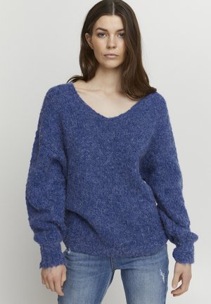 Pull en V en maille bleu texturé, avec des manches ballon et des poignets côtelés. Associé à un jean bleu clair.