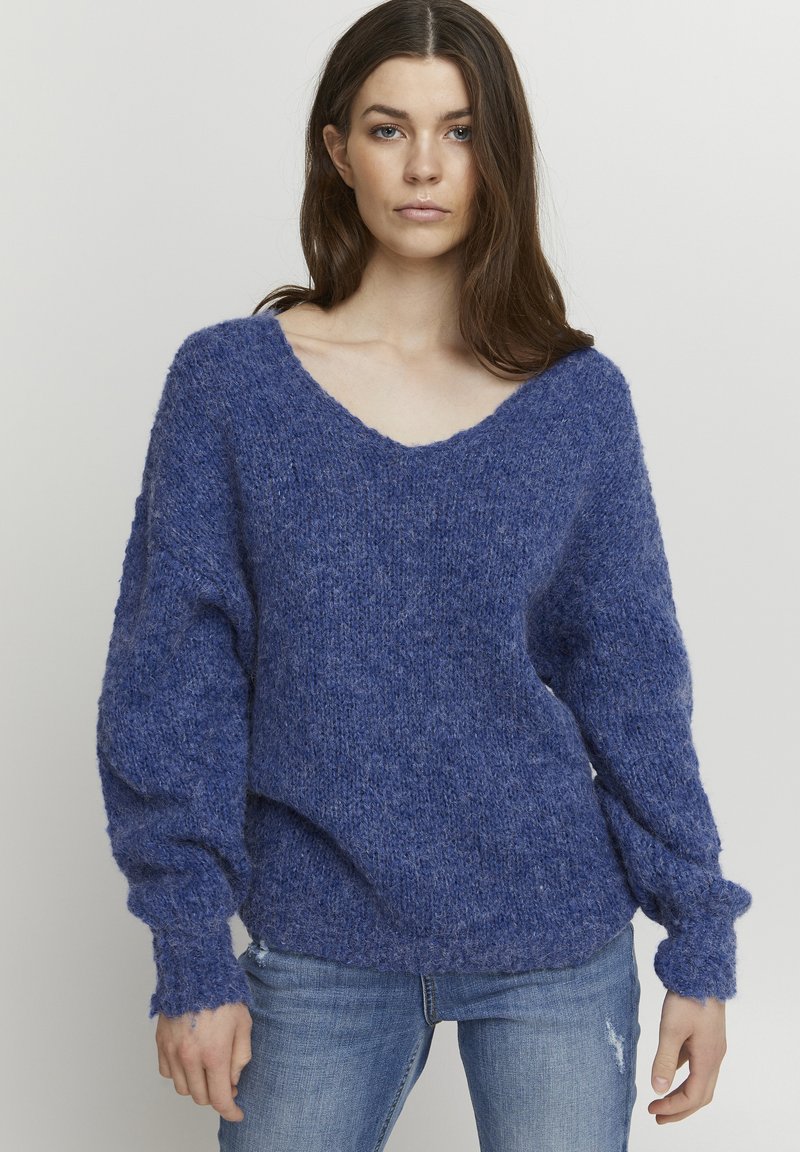 Pull en V en maille bleu texturé, avec des manches ballon et des poignets côtelés. Associé à un jean bleu clair.