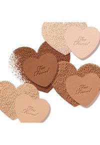Six heart-shaped pressed powders in various skin tones, featuring a mix of matte and textured surfaces, embossed with "Too Faced" branding.