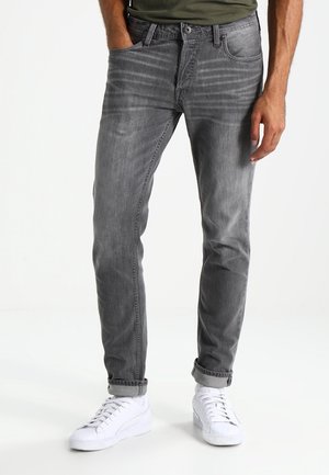 Mann trägt graue Slim-Fit-Jeans mit aufgerollten Bündchen und weißen Sneakern, steht mit einer Hand in der Tasche vor einem schlichten weißen Hintergrund.