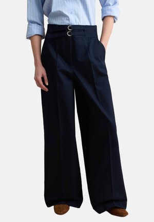 Femme portant un pantalon noir taille haute à jambes larges avec détail à double boucle, associé à une chemise rayée bleu clair et des chaussures marron.