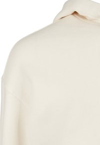 Detailný záber na pletenú látku v krémovej farbe, zobrazujúci detaily ramena a hornej rukávovej časti s jemným švom a prehnutým golierom.
