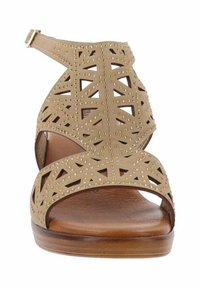 Sandalo beige con ritagli geometrici e accenti di borchie metalliche, cinturino regolabile alla caviglia, suola piatta e finitura in legno liscia.