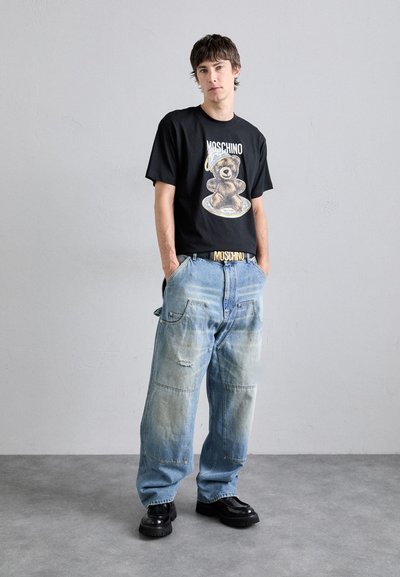 T-shirt noir à manches courtes avec un motif d'ours, associé à un jean baggy en denim bleu clair déchiré et des chaussures noires à semelles épaisses.