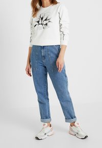 Sudadera blanca con diseño gráfico negro y costuras en contraste, combinada con vaqueros de tiro alto de mezclilla azul claro y zapatillas blancas con acentos en tonos pastel.