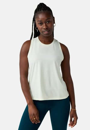 Mujer con cabello trenzado, vestida con una blusa blanca sin mangas y leggings color verde azulado, posando contra un fondo liso y claro.