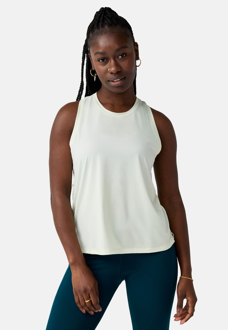 Femme aux cheveux tressés portant un haut sans manches blanc et un legging bleu sarcelle, debout devant un fond clair uni.