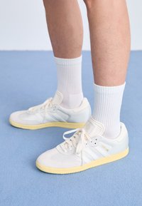 White athletic shoes with a textured mesh upper, suede accents, three white stripes, and a yellow rubber sole. Worn with white ribbed socks.