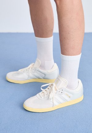 Zapatillas deportivas blancas con una parte superior de malla texturizada, detalles de ante, tres franjas blancas y una suela de goma amarilla. Llevadas con calcetines blancos de canalé.