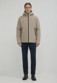 Giacca beige con cappuccio e zip frontale, dettagli neri, due tasche laterali e una texture liscia, abbinata a pantaloni blu scuro e scarpe nere.