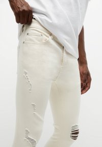 Jeans de denim blanco con detalles desgastados y rotos, diseño ajustado y un bolsillo plano con herrajes metálicos. Textura suave.