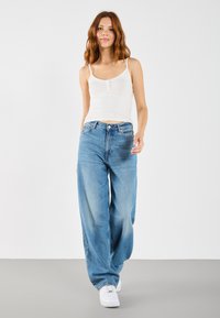 Let vægt hvid tanktop parret med løstsiddende, mellemmørke blå jeans og hvide sneakers, sat mod en ensfarvet grå baggrund.