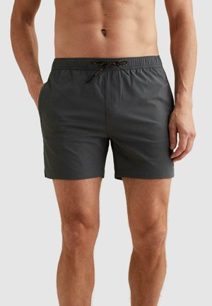 Muž nosi tmavosivé plavecké šortky s bočnými vreckami a čierno-oranžovou šnúrkou, stojaci pred jednofarebným pozadím.