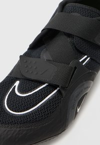 Chaussure de sport noire avec une tige en mesh, dotée d'une large sangle Velcro, d'accents texturés et d'une semelle en caoutchouc avec des détails blancs et un marquage.