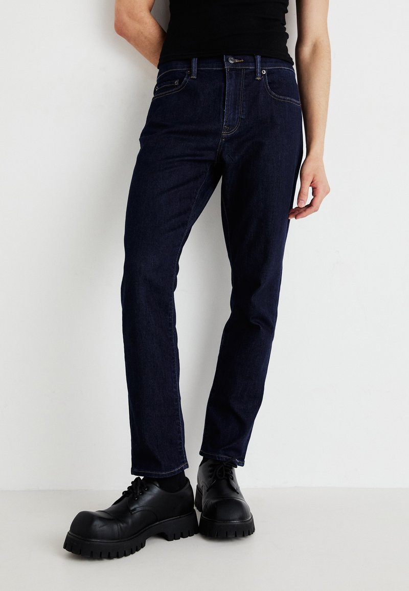 Tmavomodré džínsy, slim fit, s klasickým dizajnom piatich vreciek, jemným prešitím a zapínaním na gombík. Kombinované s robustnou čiernou obuvou.