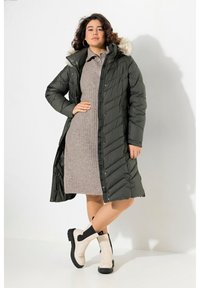 Lång olivgrön puffer-jacka med en pälsfoderhuva, tillsammans med en grå ribbad klänning och vita ankelstövlar med svarta detaljer.