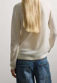 Persona con lunghi capelli biondi, vestita con un maglione crema e jeans in denim blu, in piedi contro un muro chiaro e semplice, con la schiena rivolta verso l'osservatore.