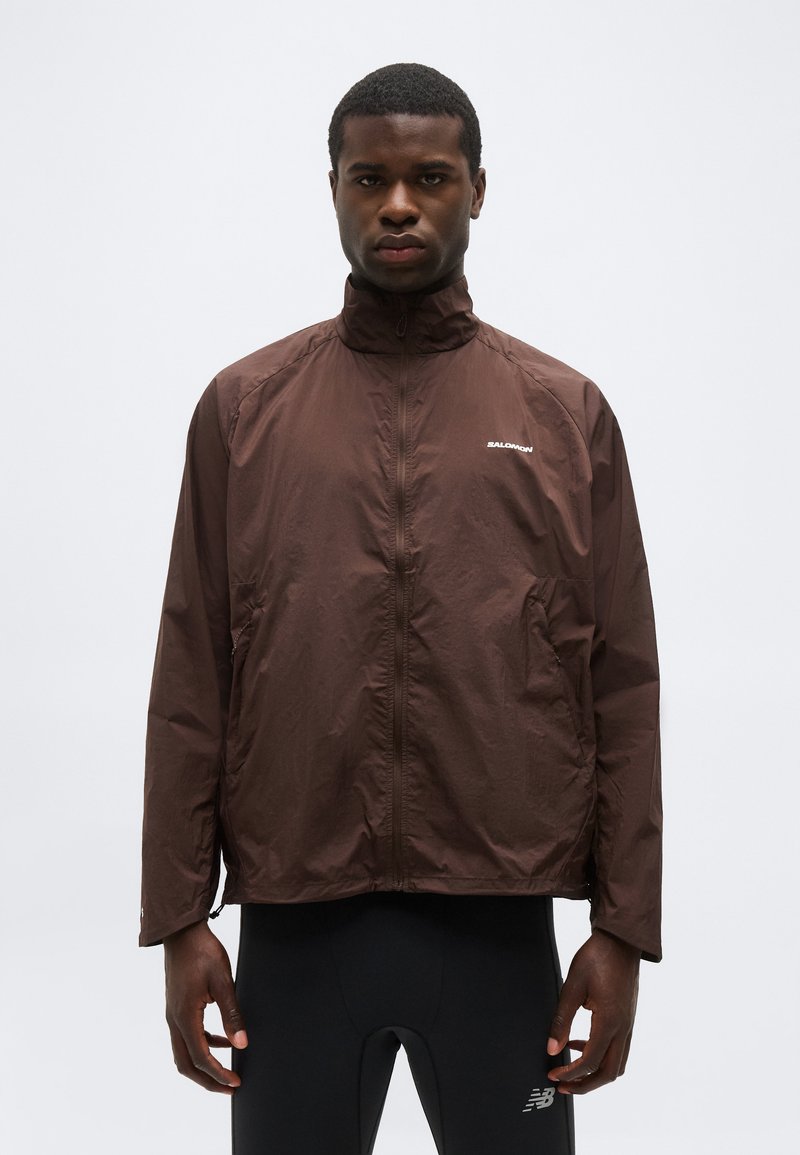 Άνδρας που φοράει καφέ ζακέτα Salomon με φερμουάρ και ψηλό γιακά, καθώς και μαύρο αθλητικό παντελόνι, πάνω σε απλό ανοιχτό φόντο.