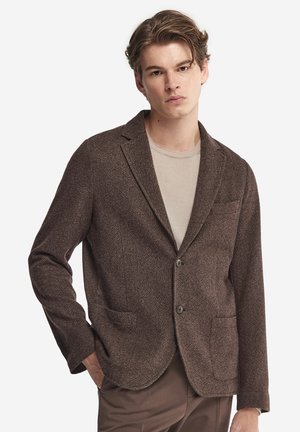 Ung mand iført en tekstureret brun blazer over en beige skjorte, med højre hånd delvist i lommen, stående foran en neutral baggrund.