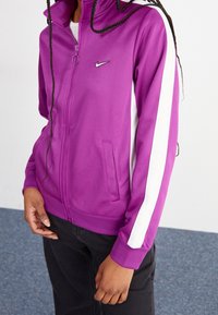 Lila Nike jacka med dragkedja och vita sidostrippar, med hög krage, två sidofickor och en slät, stretchig tygstruktur.