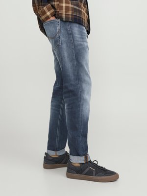 Jeans de mezclilla azul oscuro con corte recto, ligera decoloración y puños enrollados. Combinados con zapatillas negras con detalles en marrón y tela texturizada.