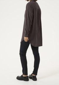 Chemise à manches longues, gris foncé, avec une coupe décontractée, présentant un ourlet arrondi et des poignets à boutons, accompagnée d'un jean skinny noir et de chaussures noires.