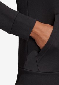 Tissu de sweat-shirt noir avec une poche avant et un poignet visible. Le matériau est lisse avec une légère élasticité et une coupe ajustée.