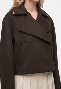 Femme portant un manteau court en laine marron foncé avec de larges revers et des poches latérales sur une jupe plissée claire.