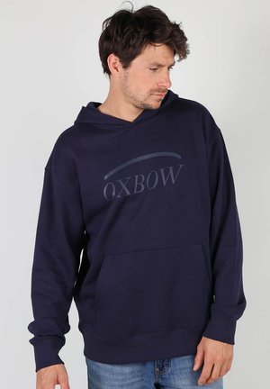 Sweat à capuche bleu marine en coton avec une poche avant, arborant un logo discret sur la poitrine. Conçu avec une coupe décontractée et une capuche.