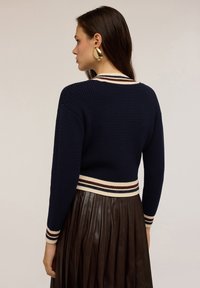 Námořnický pletený svetr s proužkovaným lemem v krémové a bordó barvě, zkrácený střih a strukturovaný povrch, spárovaný s skladanou hnědou sukní.