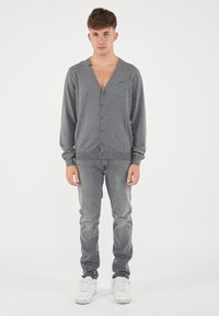 Cardigan grigio con scollo a V, maniche lunghe e chiusura con bottoni. Indossato su jeans grigi e sneakers bianche. Texture a maglia semplice e dettaglio del logo discreto.