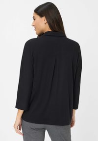 Zwarte, langmouwige top met een kraag en een plooi aan de achterkant, gemaakt van zachte stof. G gedragen met een geruite broek.