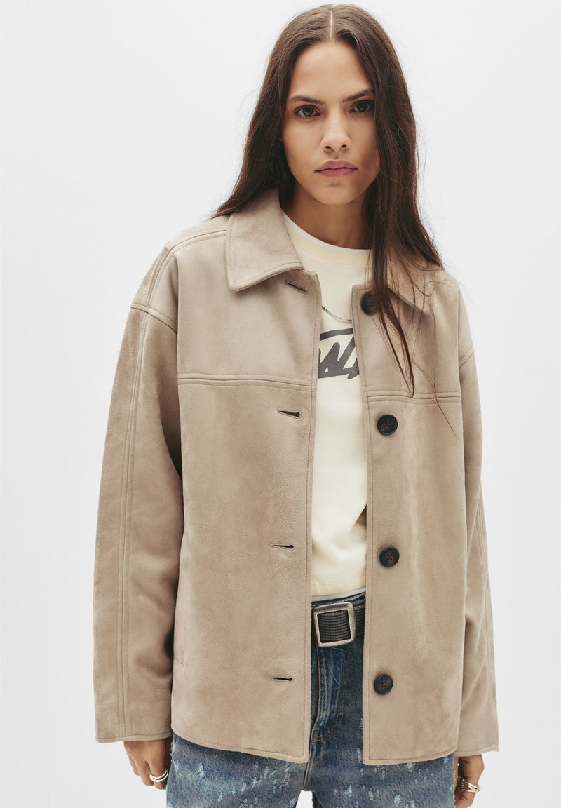 Beige suède jas met een kraag, sluiting met knopen aan de voorkant en twee grote zakken. Draag het over een crème sweatshirt en versleten denim jeans.