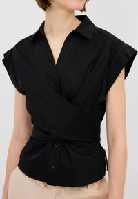 Blusa corta negra de manga corta con un cuello tipo solapa, botones sutiles y un detalle de nudo recogido en la cintura, confeccionada en una tela suave.